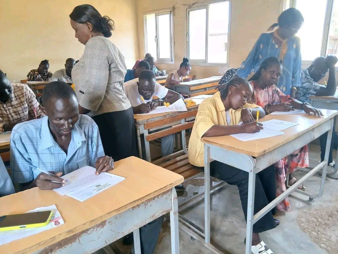 Learners write at classroom desks with two adults standing nearby